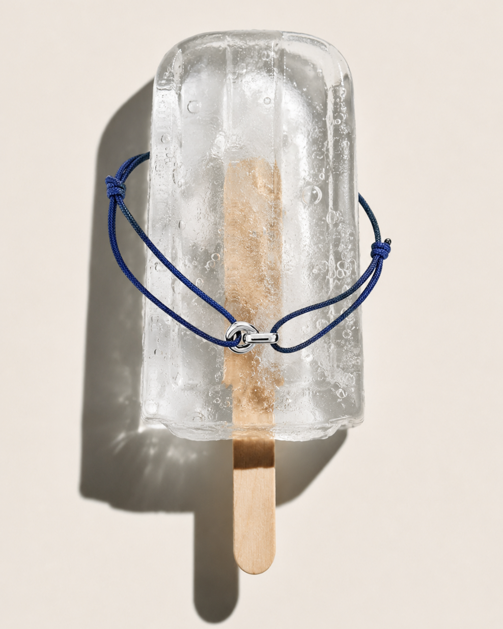 A clear ice pops mold or frozen popsicle form with a wooden stick inserted, wrapped with a dark blue cord bracelet and a small silver clasp, photographed on a warm beige background with a hard shadow.