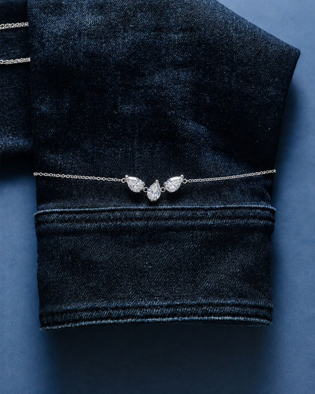 A delicate silver chain bracelet with three clear pear-cut gemstones arranged in a fan shape, displayed on a folded dark navy velvet fabric against a dark blue background.